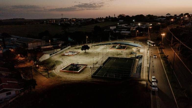 Praça Daniel Folle iluminada ao anoitecer, com quadra, pista e parquinho